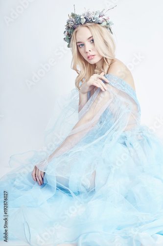 Blonde woman with wreath on her head and a gentle light blue transparent dress on a white background. Big blue eyes and beautiful skin. Fabulous mysterious magical image of a woman