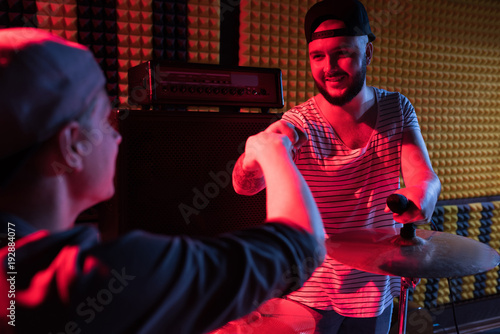 Portrait of tattooed modern singer fitstbumping drummer while rehearsing in dim recording studio photo