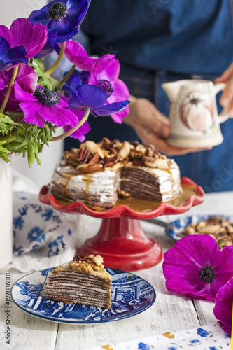 A woman and a cake of thin pancakes with white cream and nuts. Milkman with flowers in the hands of the girl. Spring fresh flowers. Breakfast in the morning. Card.