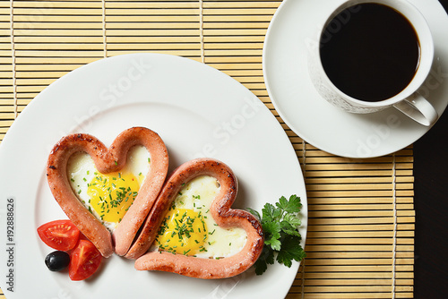 Scrambled eggs in the form of heart on a white plate with sausages, tomatoes, greens and coffee with lemon.
