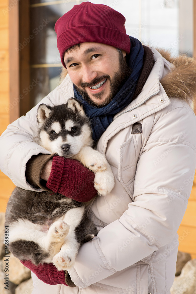 Portrait of cheerful Asian man holding puppy looking at camera and smiling happily while posing outdoors on winter day
