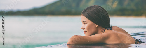 Luxury vacation woman relaxing in infinity swimming pool on summer travel at beach resort. Asian girl tourist on wellness spa relaxation outside in nature landscape banner panorama.