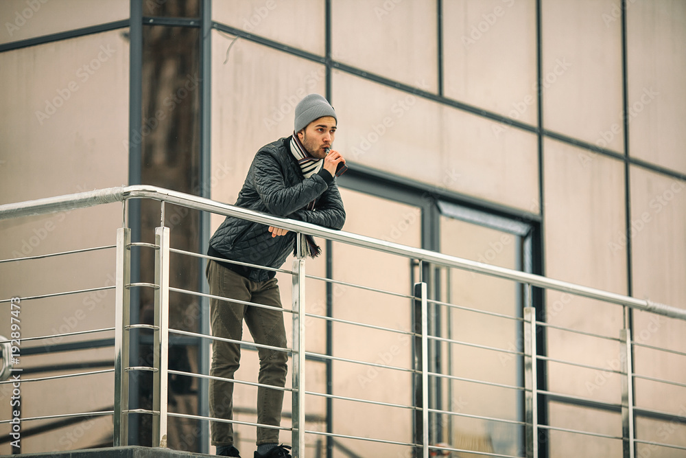 Vape man. Portrait of a handsome young white guy in casual clothes vaping an electronic cigarette opposite the futuristic modern building. Lifestyle.