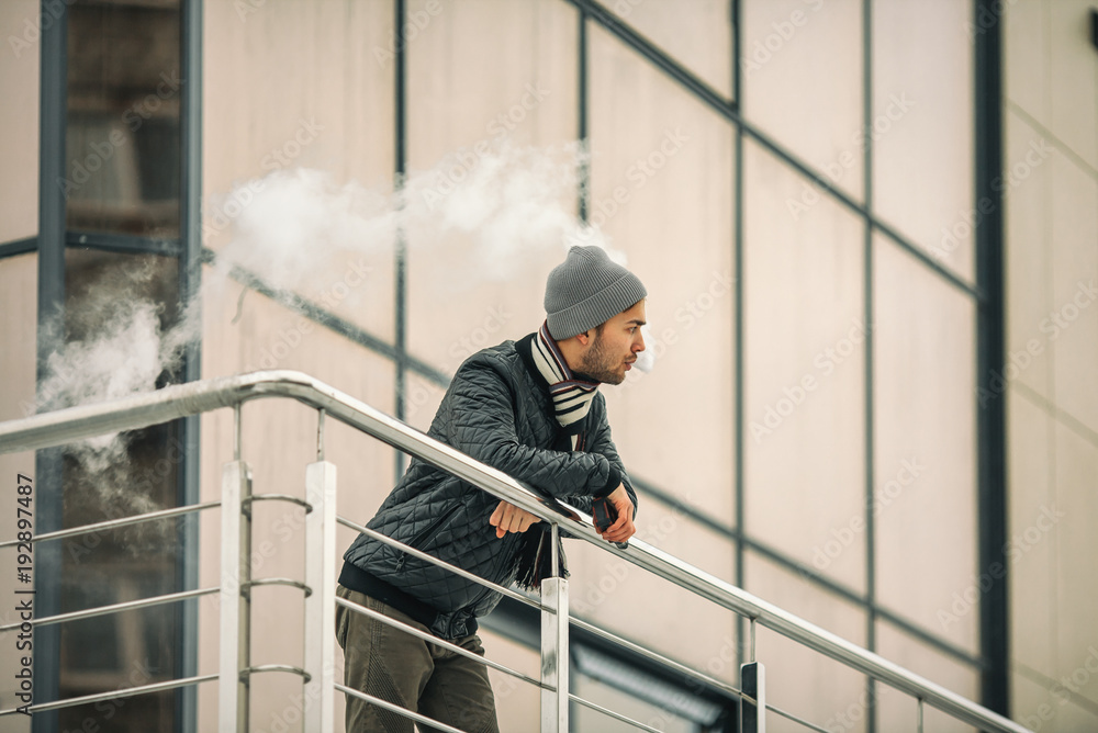 Vape man. Portrait of a handsome young white guy in casual clothes vaping an electronic cigarette and letting out puffs of steam opposite the futuristic modern building. Lifestyle. Close up.