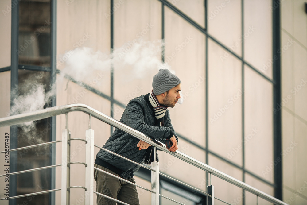 Vape man. Portrait of a handsome young white guy in casual clothes vaping an electronic cigarette and letting out puffs of steam opposite the futuristic modern building. Lifestyle. Close up. Vape man. Portrait of a handsome young white guy in casual clothes vaping an electronic cigarette and letting out puffs of steam opposite the futuristic modern building. Lifestyle. Close up.