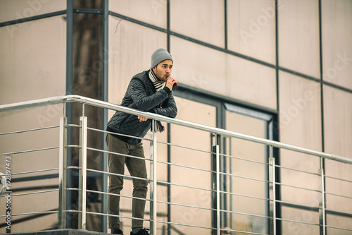 Vape man. Portrait of a handsome young white guy in casual clothes vaping an electronic cigarette opposite the futuristic modern building. Lifestyle. © aleksandr_yu