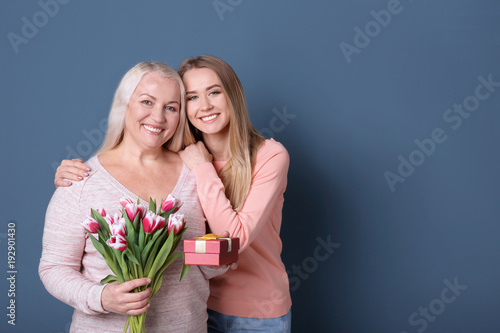 Young daughter with mother holding bouquet of flowers and gift box on color background