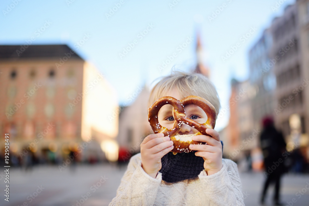 Obraz premium Little tourist holding traditional bavarian bread called pretzel on the town hall building background in Munich, Germany