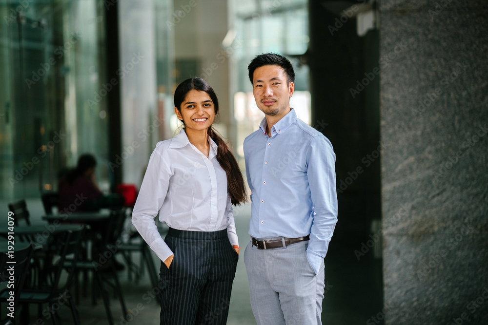 Portrait of two colleagues standing in their office during day. One is an Indian woman, and the other a Chinese man. They are smiling in a relaxed way. Portrait of two colleagues standing in their office during day. One is an Indian woman, and the other a Chinese man. They are smiling in a relaxed way.