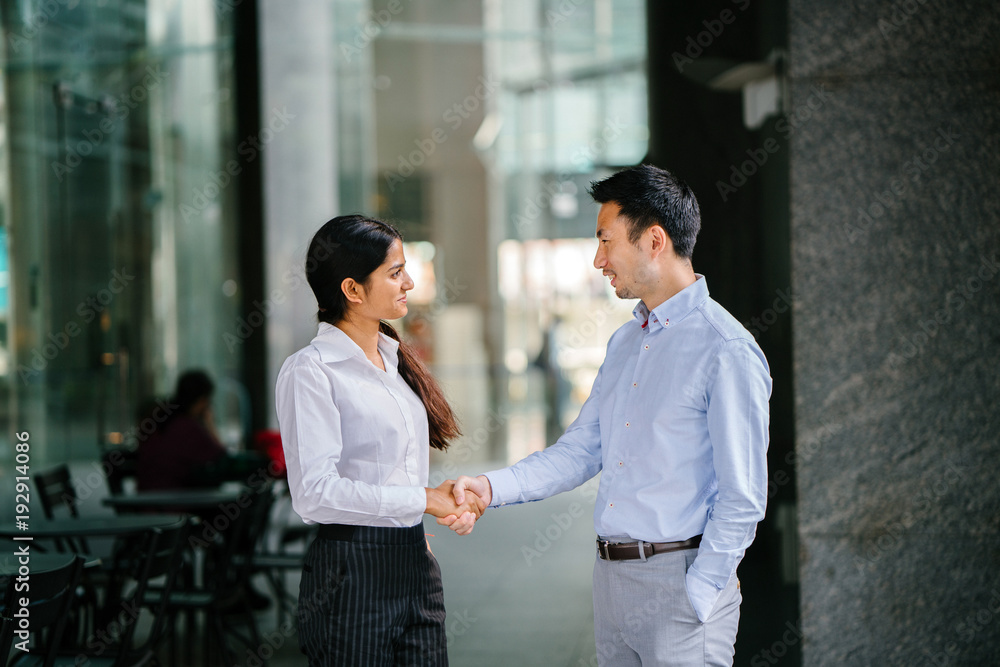 An image of two colleagues who shake hands for success while standing in their office during the day. They smile in a relaxed manner and wear both a professional outfit. An image of two colleagues who shake hands for success while standing in their office during the day. They smile in a relaxed manner and wear both a professional outfit.