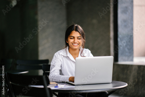 An image of a young Indian Asian woman typing and working in a laptop and searching for a job. She is wearing a professional, crisp white shirt and she is smiling as she sits at a table during the day