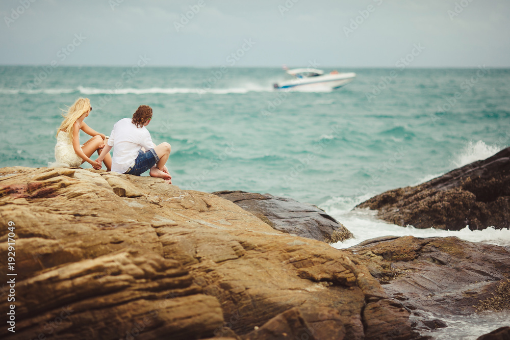 couple in love sit on rocky beach. newlyweds looking at the passing boat.