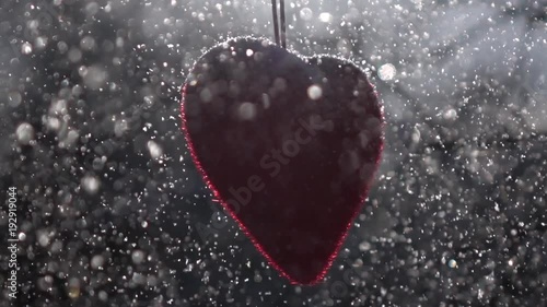 Winter lyric scene of Saint Valentine's Day. Red heart, hanging in back light, and snow mass, falling on it. Love Day of winter cold February. Cold sunny weather for hot romantic heart on forest.