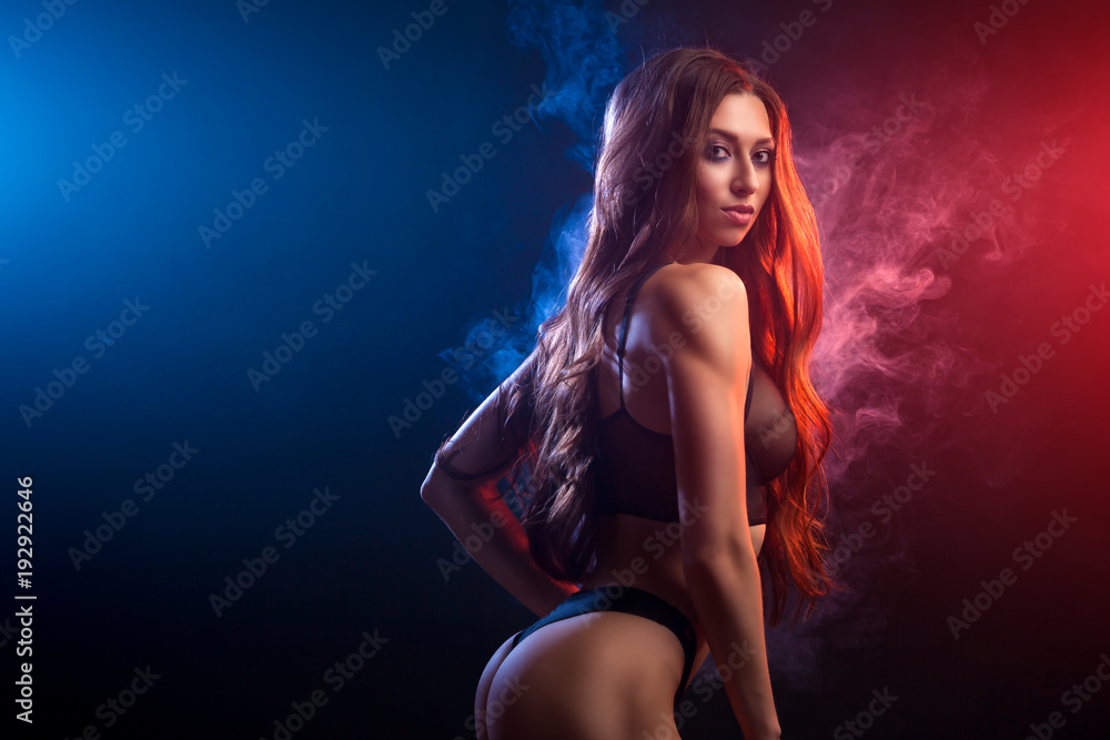 Young dark-haired woman in black lingerie posing against a background of red and   blue smoke from a vape on a black isolated background
