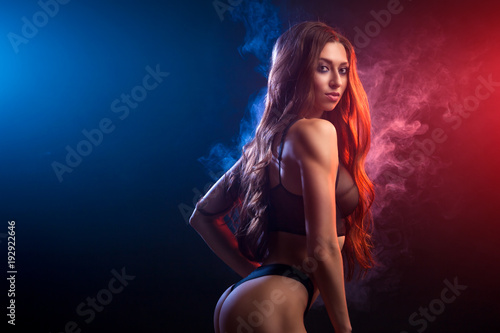 Young dark-haired woman in black lingerie posing against a background of red and blue smoke from a vape on a black isolated background