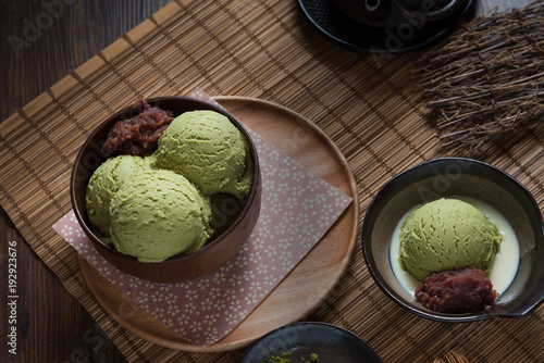 Green tea matcha ice cream scoops with sweet red bean paste in dark brown bowl over wooden plate with green tea powder on bamboo plate mat on wood table, Japanese style sweet, flat lay (top view)