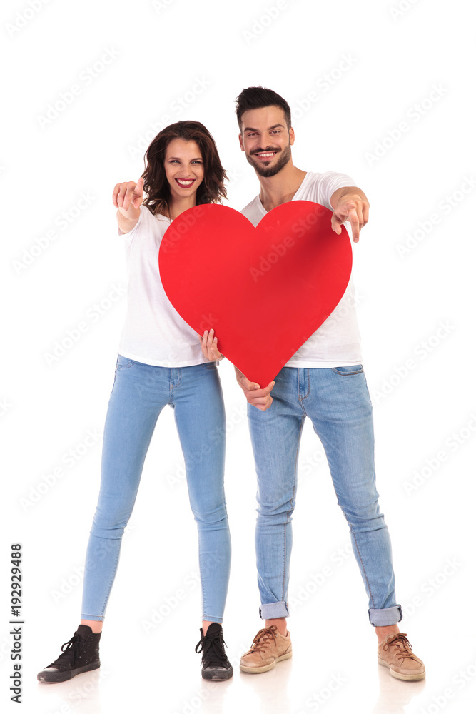 young casual couple holding a big heart and points fingers