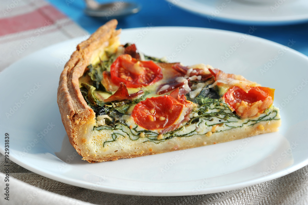 A piece of classic French pie quiche on a white plate. The composition of the filling is spinach, bacon, cheese, cherry tomatoes. The background is draped with a napkin. Close-up. Macro photography. A piece of classic French pie quiche on a white plate. The composition of the filling is spinach, bacon, cheese, cherry tomatoes. The background is draped with a napkin. Close-up. Macro photography.