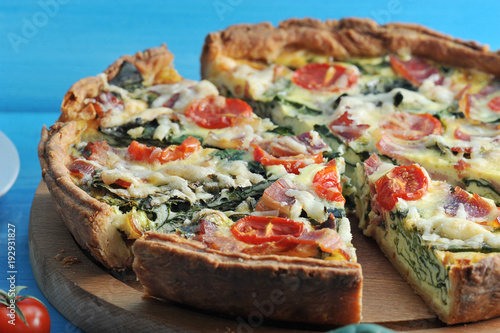 French classic pie quiche with spinach, bacon, cherry tomatoes and cheese. The pie on the wooden board is cut into portions. Blue background. Close-up. Macro photography.