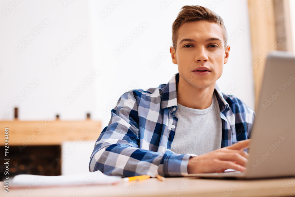 I like studying. Good-looking unsmiling fair-haired boy working on his laptop and wearing a tartan shirt while sitting at the table I like studying. Good-looking unsmiling fair-haired boy working on his laptop and wearing a tartan shirt while sitting at the table