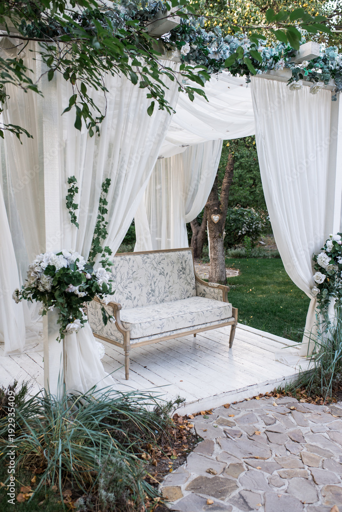 Obraz premium In the garden there is a podium on which a beautiful white sofa in the style of Provence or rustic. Above the sofa is an arch with blossoms and flowing white fabric. Decoration for a wedding, scenery