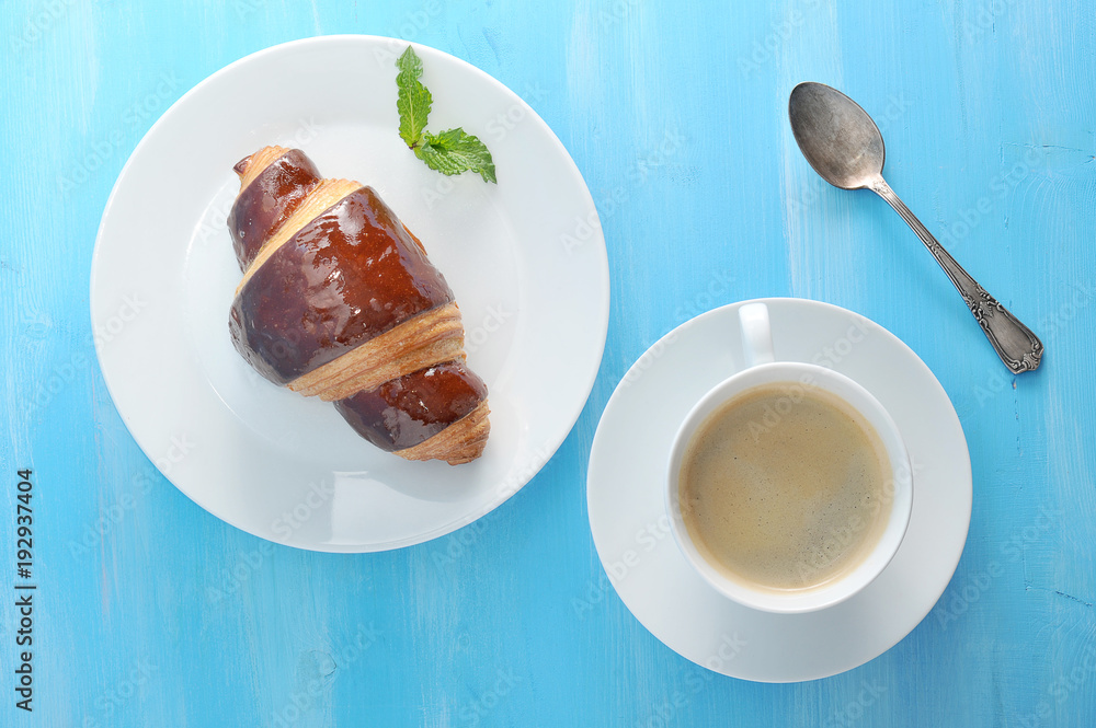 A lush and airy chocolate croissant and a cup of black coffee are the perfect combination for breakfast or a snack. On a plate with a croissant a mint leaf. Blue wood background. View from above. A lush and airy chocolate croissant and a cup of black coffee are the perfect combination for breakfast or a snack. On a plate with a croissant a mint leaf. Blue wood background. View from above.