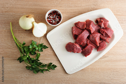 A set for cooking minced meat. Pieces of beef on a white tray. Next to onions, parsley and a cup of pepper and salt. Close-up. Light wooden background.