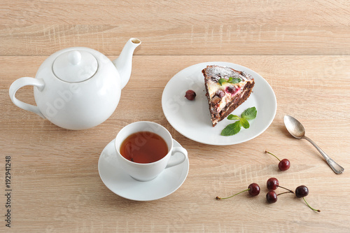 Classic chocolate brownie on a plate. Brownie is decorated with a leaf of mint. Next cup with tea and a teapot. Fresh cherries are on the table. Serving for one person. Light wooden background.