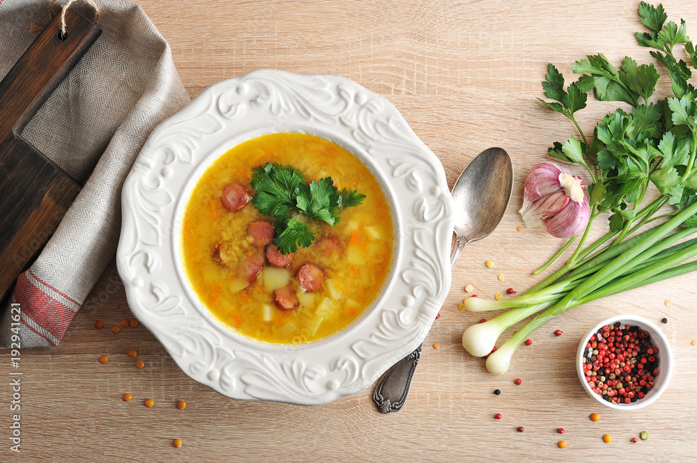 Pea soup with smoked meat in a plate. Fabric napkin. Next to the plate, a spoon and spices: garlic, parsley, green onions and peppers. Light wooden background. View from above. Pea soup with smoked meat in a plate. Fabric napkin. Next to the plate, a spoon and spices: garlic, parsley, green onions and peppers. Light wooden background. View from above.