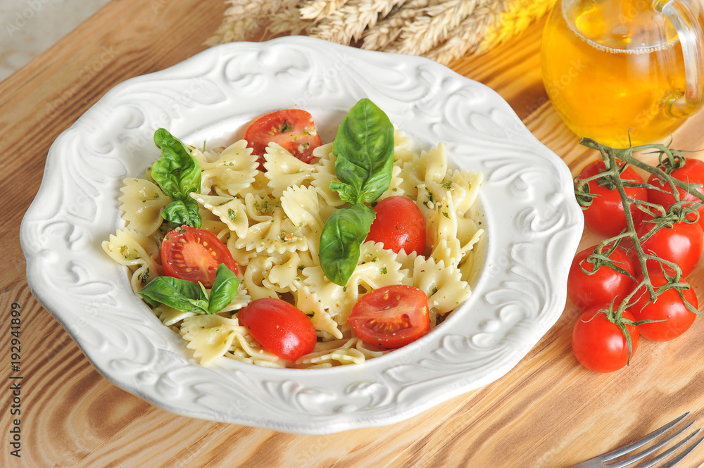 Pasta with pesto sauce and cherry tomatoes. The pasta is decorated with fresh basil leaves. Next to the dish is a jug of olive oil and a twig of tomato. Light wooden background. Macro photography. Pasta with pesto sauce and cherry tomatoes. The pasta is decorated with fresh basil leaves. Next to the dish is a jug of olive oil and a twig of tomato. Light wooden background. Macro photography.