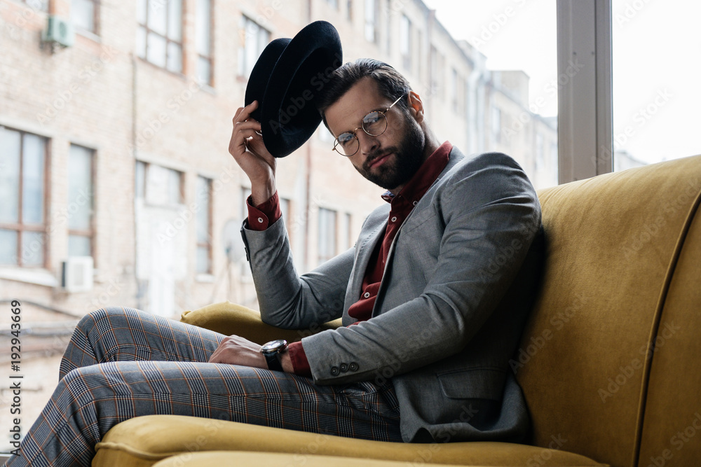 fashionable elegant man in eyeglasses and hat sitting on couch
