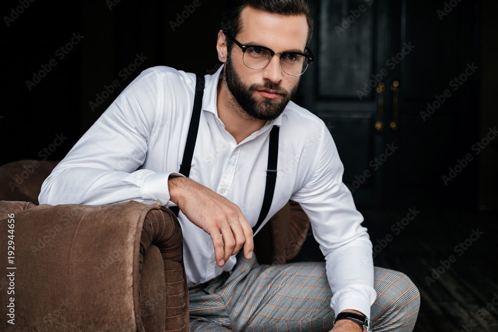 handsome elegant man in eyeglasses, white shirt and suspenders sitting in armchair handsome elegant man in eyeglasses, white shirt and suspenders sitting in armchair