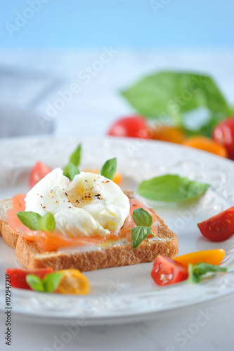 Toast with salted salmon and egg poached on a plate. The dish is decorated with multi-colored cherry tomatoes and basil leaves. Light background. Close-up. Vertical orientation of the frame.