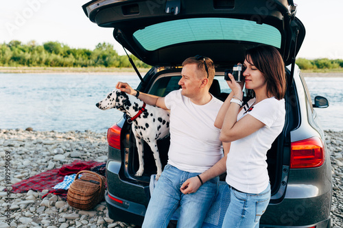 Portrait of happy young adult couple with dog on roadtrip. Man sitting on plaid with woman. Outdoor picnic concept.