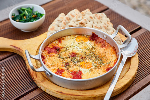 Scrambled eggs with tomatoes, cheese and spices in an aluminum frying pan with handles with homemade thin lavash and juicy greens on a wooden brown table. The photo was taken under natural light.