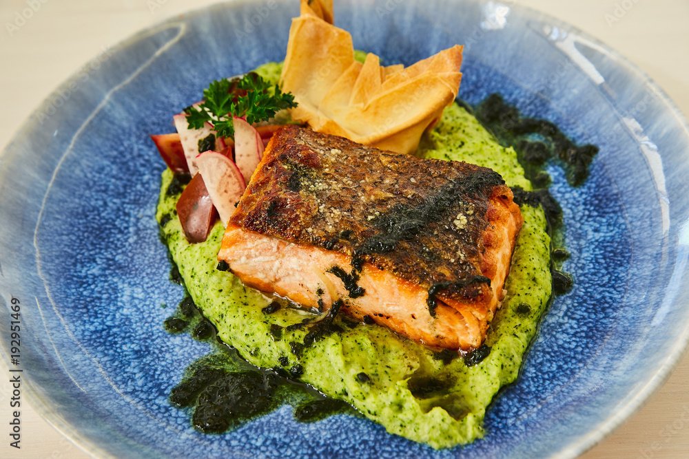 Large juicy roasted fatty piece of salmon steak on a cushion of vegetable puree, decorated with radish, parsley, cilantro and baked dough. Fish dish in a beautiful blue plate on a white wooden table.