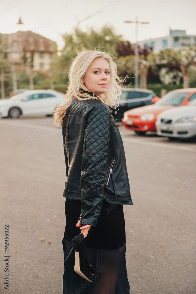 Outdoor portrait of beautiful blond woman wearing black leather jacket, holding high heel shoes, walking away