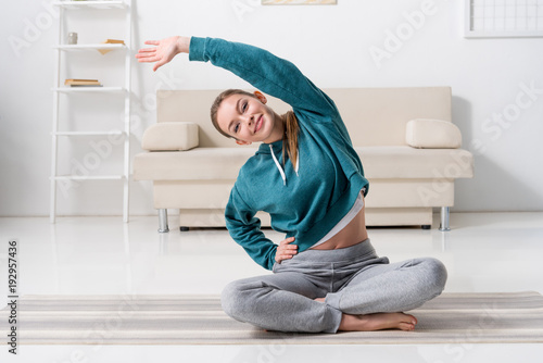 smiling girl sitting in lotus position on yoga mat and stretching at home