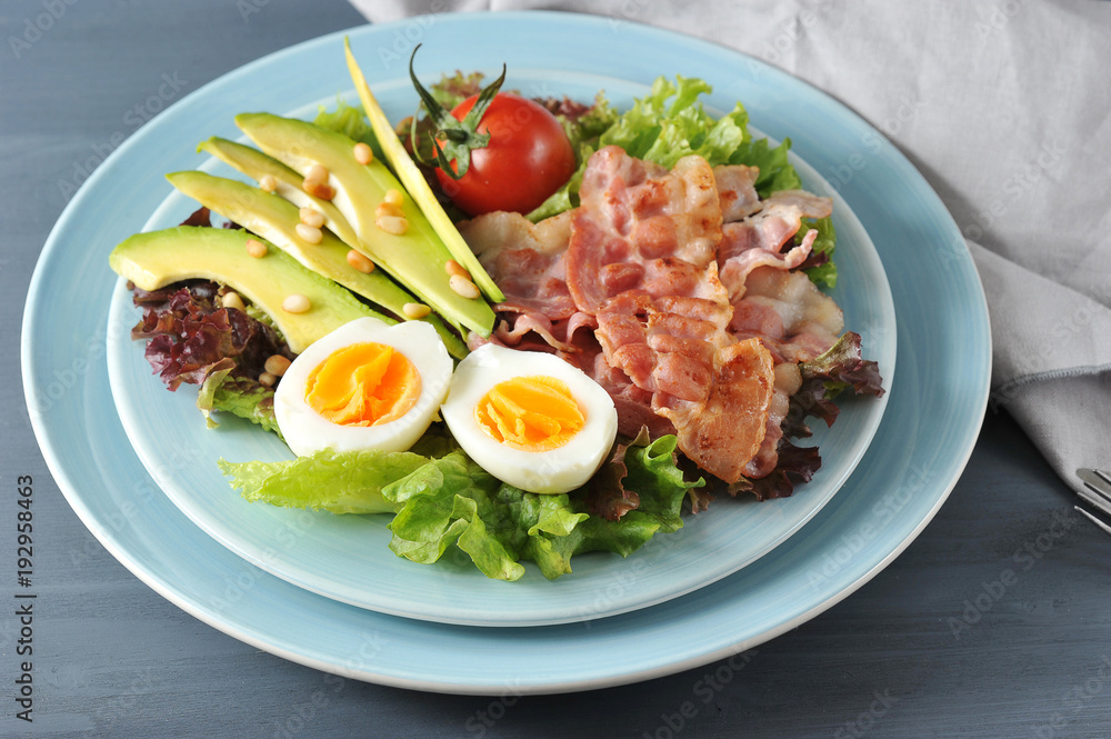 Salad with avocado and bacon. In a deep plate lettuce leaves, hard-boiled egg, fried bacon, avocado slices, tomato, pine nuts. In the picture, a napkin. Dark background. Close-up. Macro photography. Salad with avocado and bacon. In a deep plate lettuce leaves, hard-boiled egg, fried bacon, avocado slices, tomato, pine nuts. In the picture, a napkin. Dark background. Close-up. Macro photography.