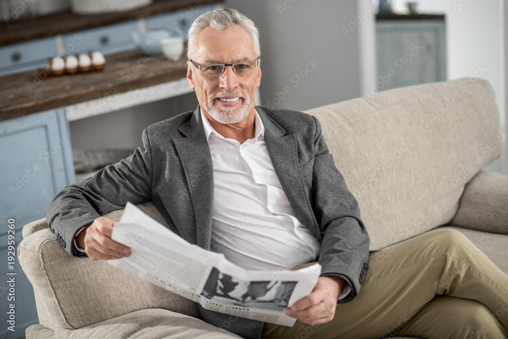 Joyful mood. Positive delighted man crossing legs while sitting on his sofa and smiling on camera Joyful mood. Positive delighted man crossing legs while sitting on his sofa and smiling on camera
