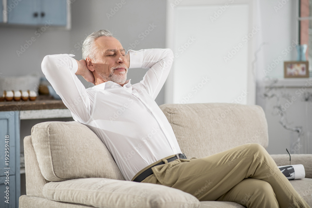 Time to dream. Positive delighted bearded man crossing legs while sitting on the sofa, stretching arms