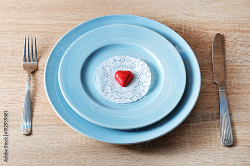 Two blue plates one on the other. A white paper napkin is on the plate. On a napkin is a red candy in the shape of a heart. There are cutlery nearby. Light wooden background. View from above. Close-up