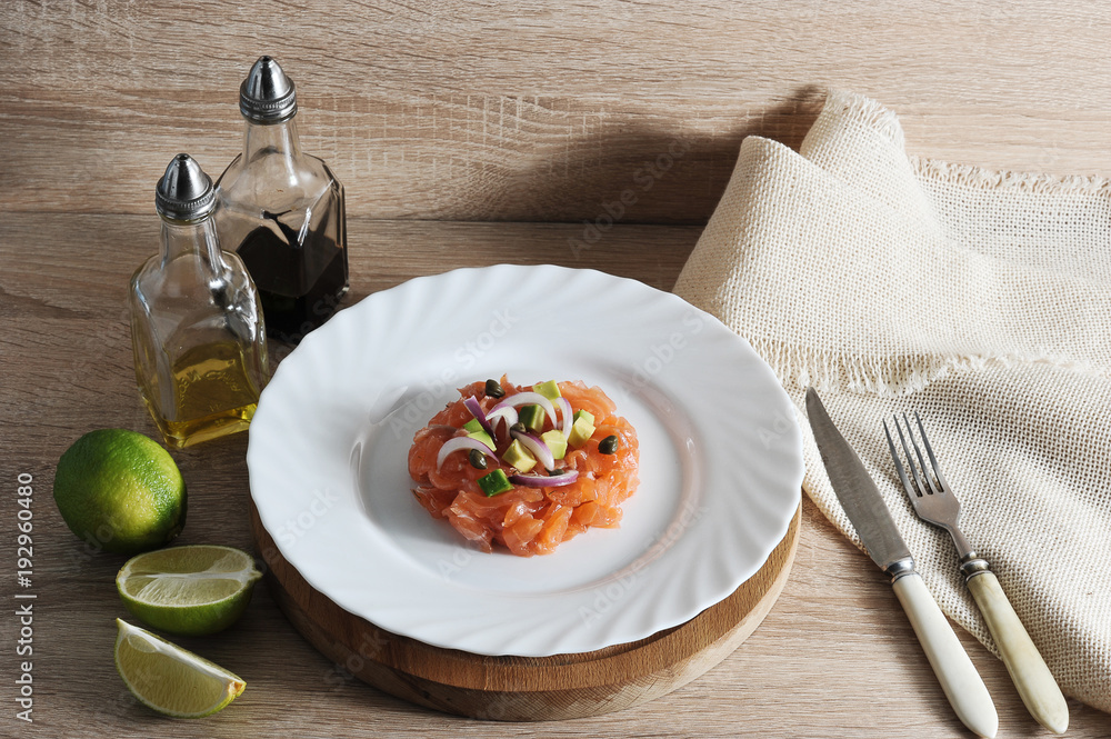 Tartar salmon on a plate. The dish is decorated with pieces of avocado, capers, red onions. In the frame of the tank with butter and balsamic sauce, lime, napkin, cutlery. Light background. Close-up. Tartar salmon on a plate. The dish is decorated with pieces of avocado, capers, red onions. In the frame of the tank with butter and balsamic sauce, lime, napkin, cutlery. Light background. Close-up.