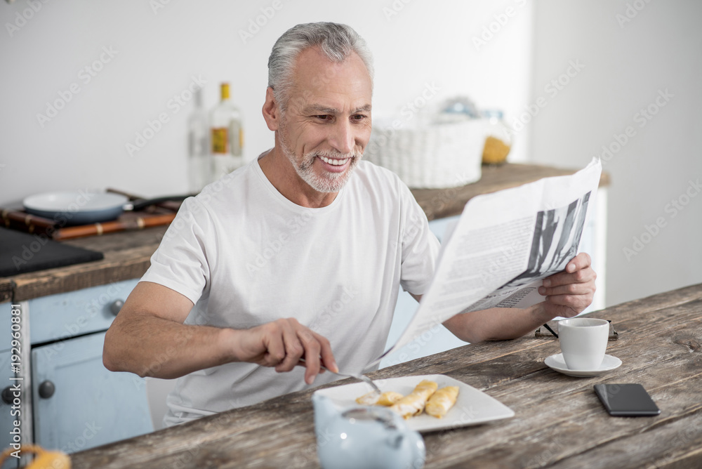 Morning routine. Happy bearded man keeping smile on his face while reading article and having breakfast Morning routine. Happy bearded man keeping smile on his face while reading article and having breakfast