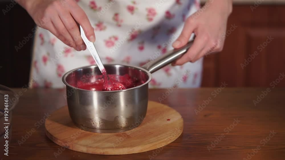 Woman is stirring jam in saucepan. She is standing near table and holding bowl, hand with spoon is mixing, close-up Woman is stirring jam in saucepan. She is standing near table and holding bowl, hand with spoon is mixing, close-up