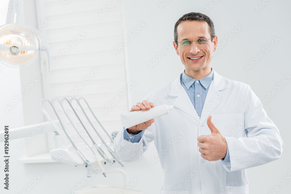 Dentist showing blank toothpaste tube and thumb up in modern dental clinic Dentist showing blank toothpaste tube and thumb up in modern dental clinic