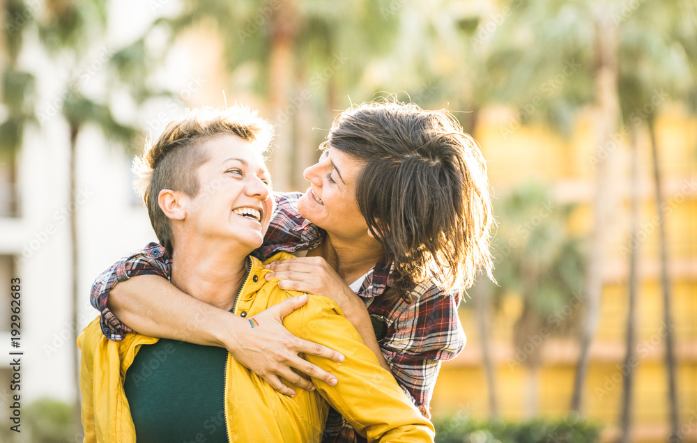 Naklejka premium Happy playful girlfriends in love sharing time together at travel trip on piggyback hug - Women friendship concept with girls couple having fun on fashion clothes outdoors - Bright warm sunset filter