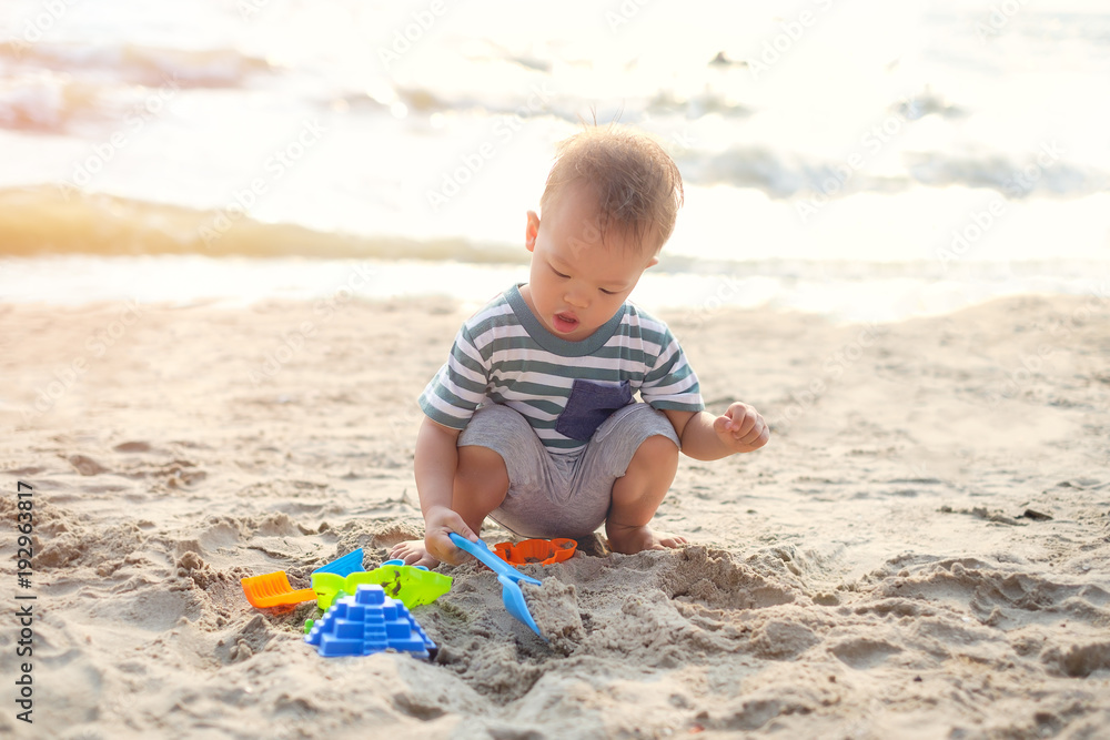 Cute little Asian 18 months, 1 years old toddler boy sitting & playing children's beach toys on beautiful sandy tropical sunset beach, Family travel, water outdoor activity on beach vacation concept Cute little Asian 18 months, 1 years old toddler boy sitting & playing children's beach toys on beautiful sandy tropical sunset beach, Family travel, water outdoor activity on beach vacation concept