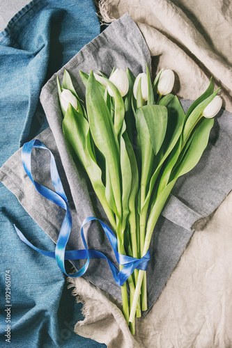 Bouquet of white spring tulips with leaves and blue ribbon over different cloth background. Top view, space.