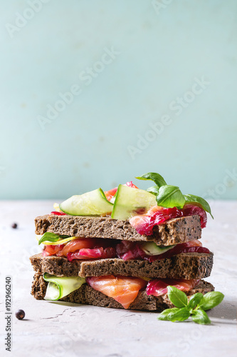 Sliced beetroot marinated salmon sandwiches with rye bread, cucumber, basil and lemon served in stack on wooden cutting board over grey blue texture background. Close, up, space